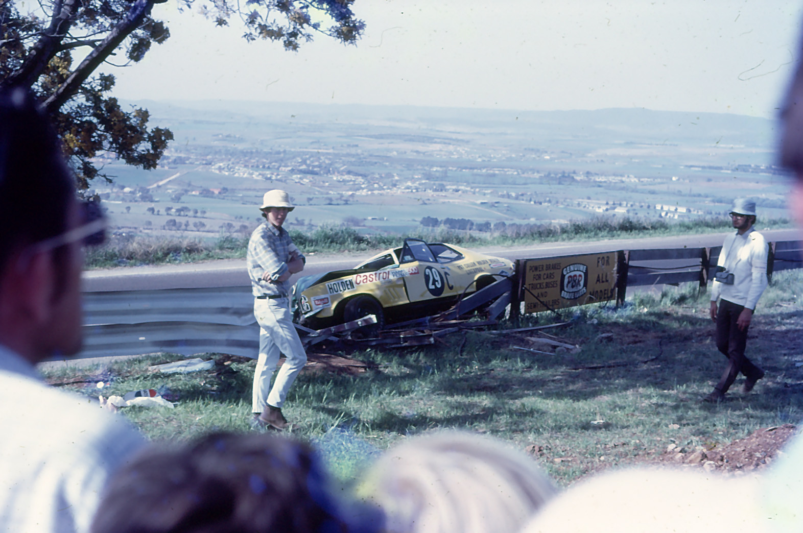 A yellow Holden racing car, number 29, rests against a damaged safety barrier following an accident at Mount Panorama, Bathurst. Spectators survey the scene, with the sweeping countryside and town of Bathurst visible in the valley below.